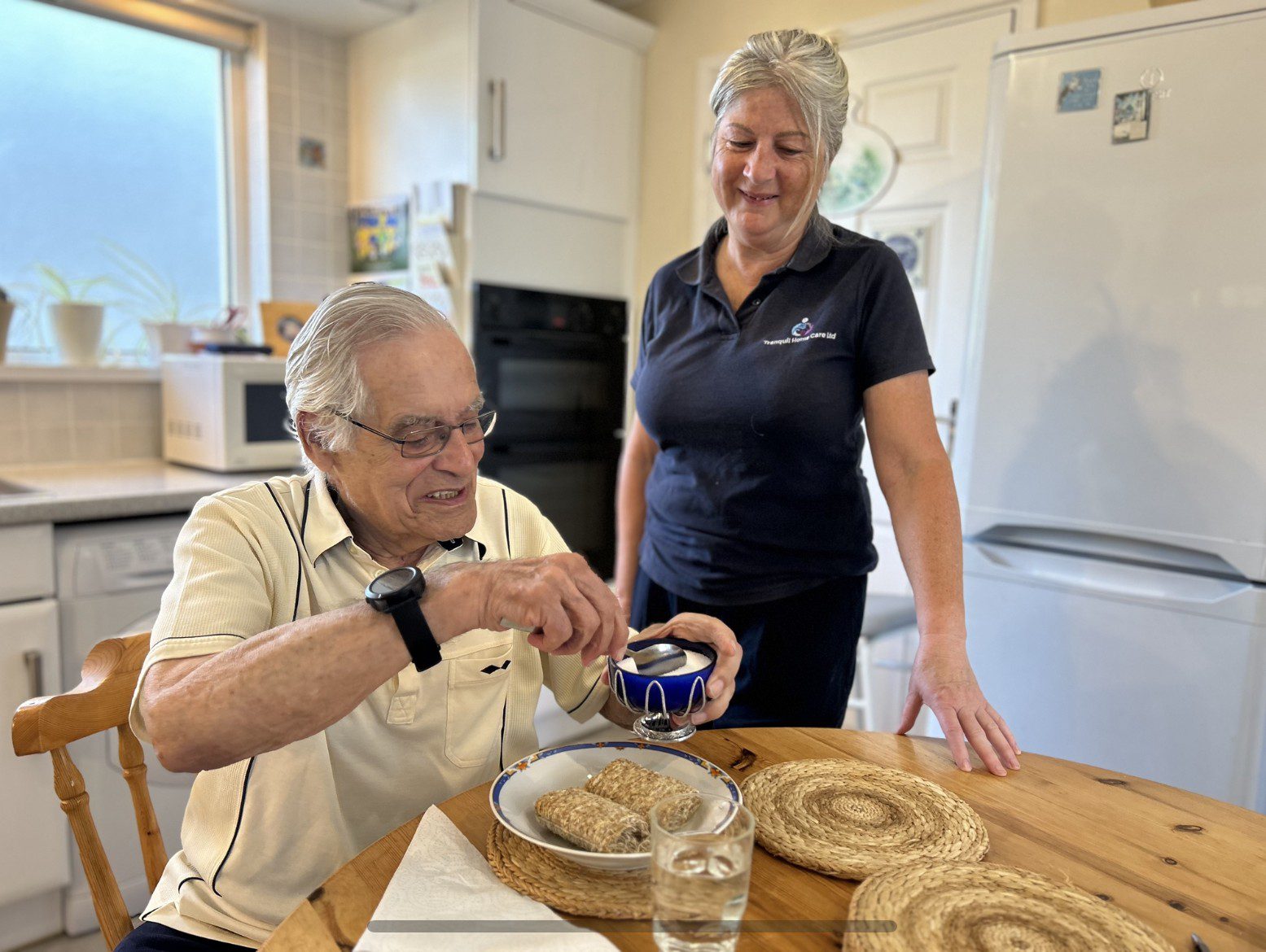 Elderly man enjoying tea with caregiver nearby.